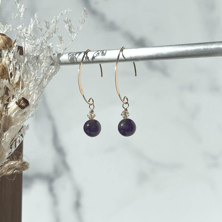 These elegant Amethyst earrings from our collection promise to make a chic statement wherever you go. The flat V-shaped half-hoop ear wire is crafted from 14Kt Rose Gold-filled metal, and each earring is adorned with a shimmery golden Swarovski crystal bead.