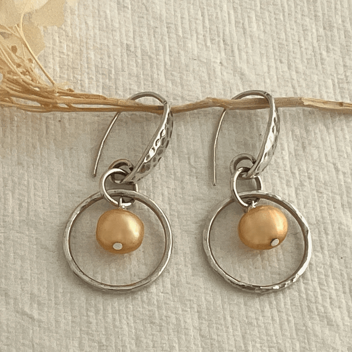 Handcrafted small sterling silver hoops adorned with apricot colored freshwater pearls and a shimmery hammered texture.  Shown on a white paper background.