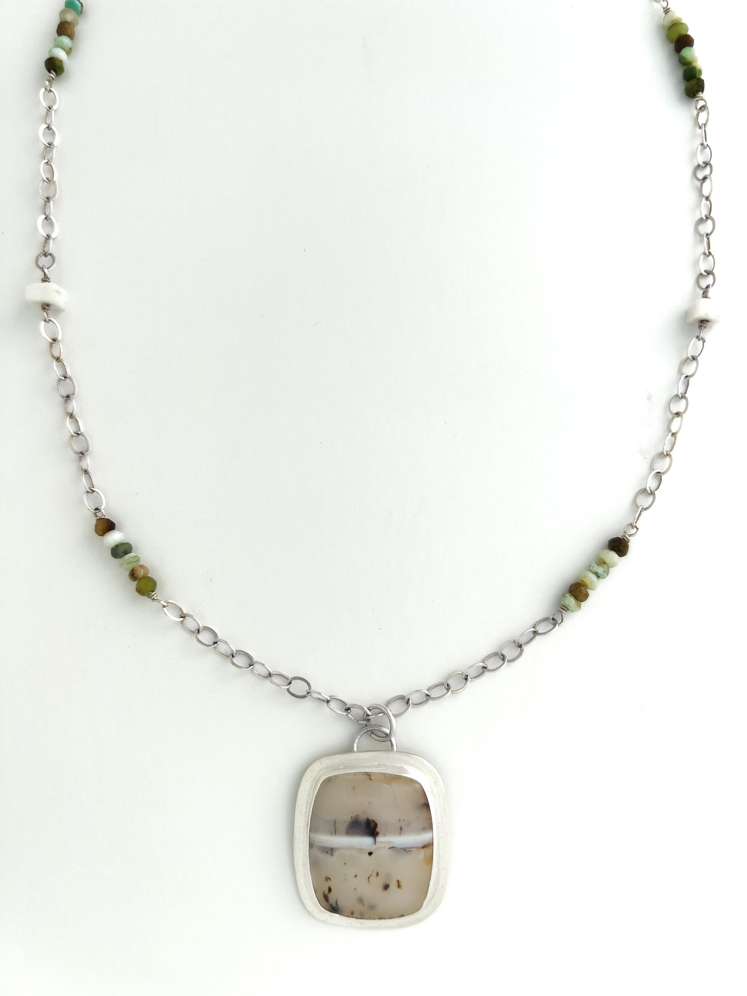 Dendritic Opals are known for their unique and intricate patterns, reminiscent of natural landscapes. This pendant necklace features an elegant setting that beautifully showcases this distinctive stone. Peruvian Blue Opals and White Moonstone beads intricately intertwine within a sterling silver chain, creating a one-of-a-kind necklace.