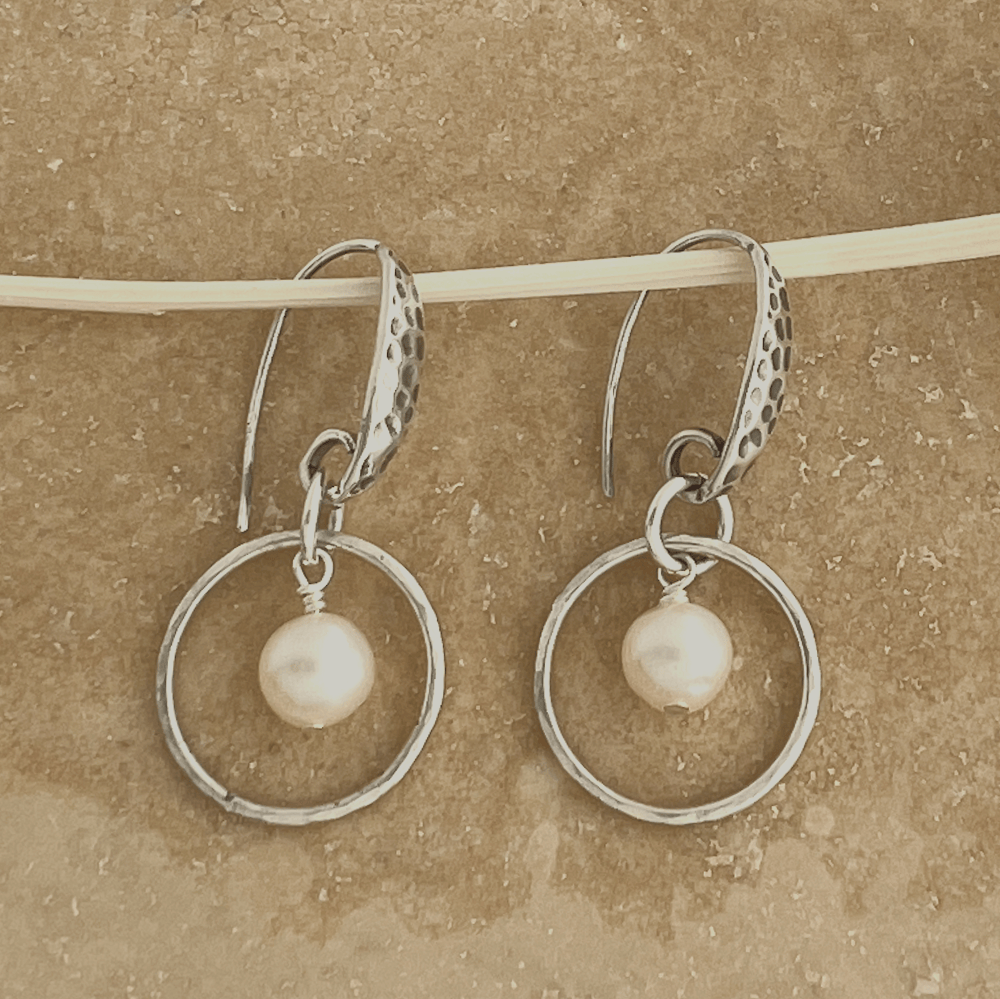 Elegantly crafted small sterling silver hoops adorned with delicate freshwater pearls and a shimmery hammered texture.  Shown on a beige background.